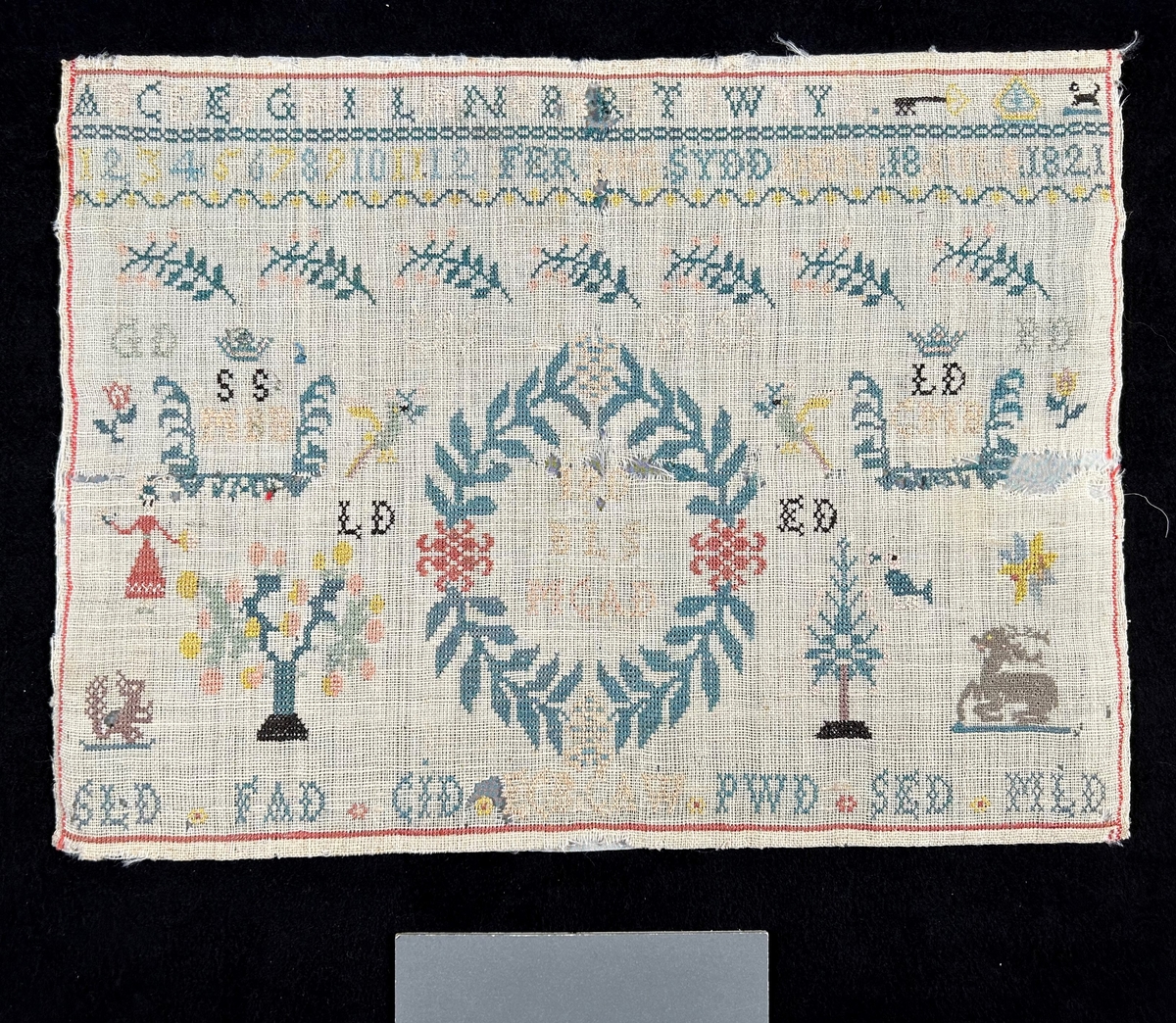 Märkduk i symmetrisk komposition. Överst två rader med alfabetet och siffror. På den undre raden står "Ferdigsydd den 18 juli 1821." I mitten en oval krans med monogram; JPD, BLLS, MCAD, på båda sior två par krönta monogram SS, MBB LD, CMB. Längst ned en rad med olika monogram; SLD, FAD, CJD, ECB, CAW, PWD, SED, MLD. För övrigt växt- och djurmotiv. Monterad på papper.