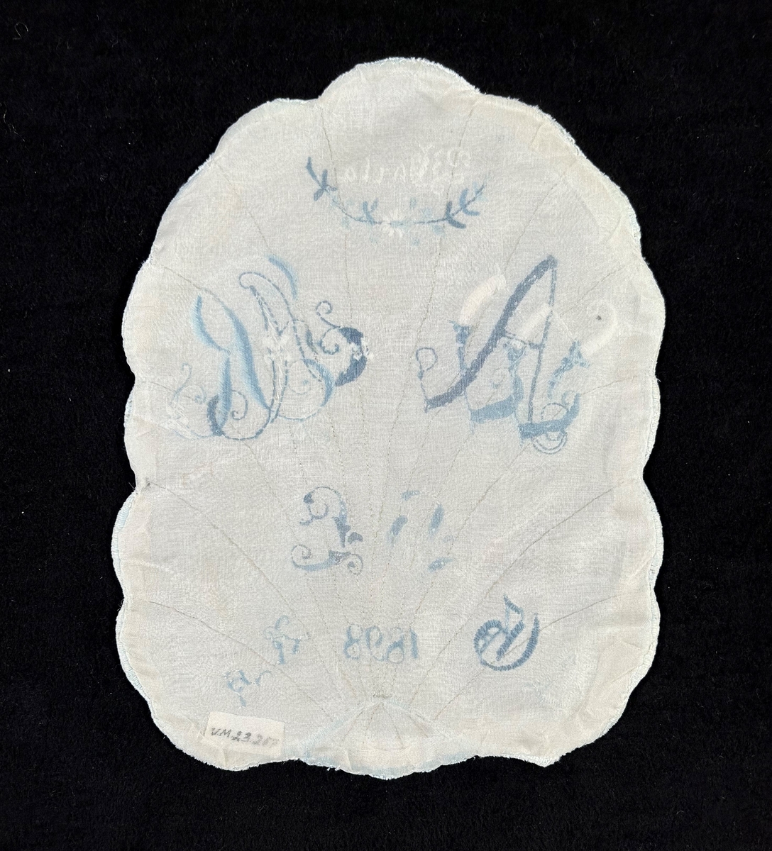 Märkduk av vitt linne med formen av ett blad med nerver av beige silke i stjälksöm. Allt märkt i blått och vitt märkgarn. Vit langett runt om.