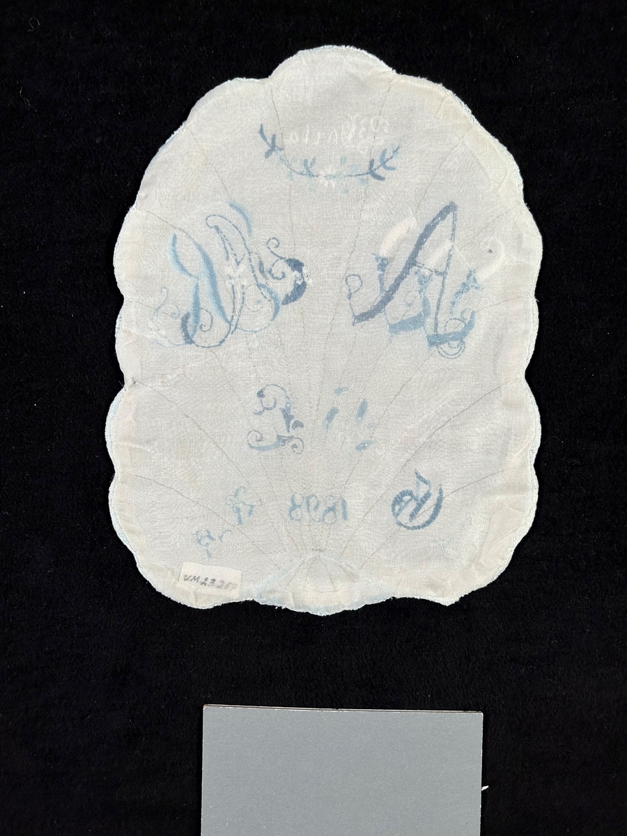 Märkduk av vitt linne med formen av ett blad med nerver av beige silke i stjälksöm. Allt märkt i blått och vitt märkgarn. Vit langett runt om.