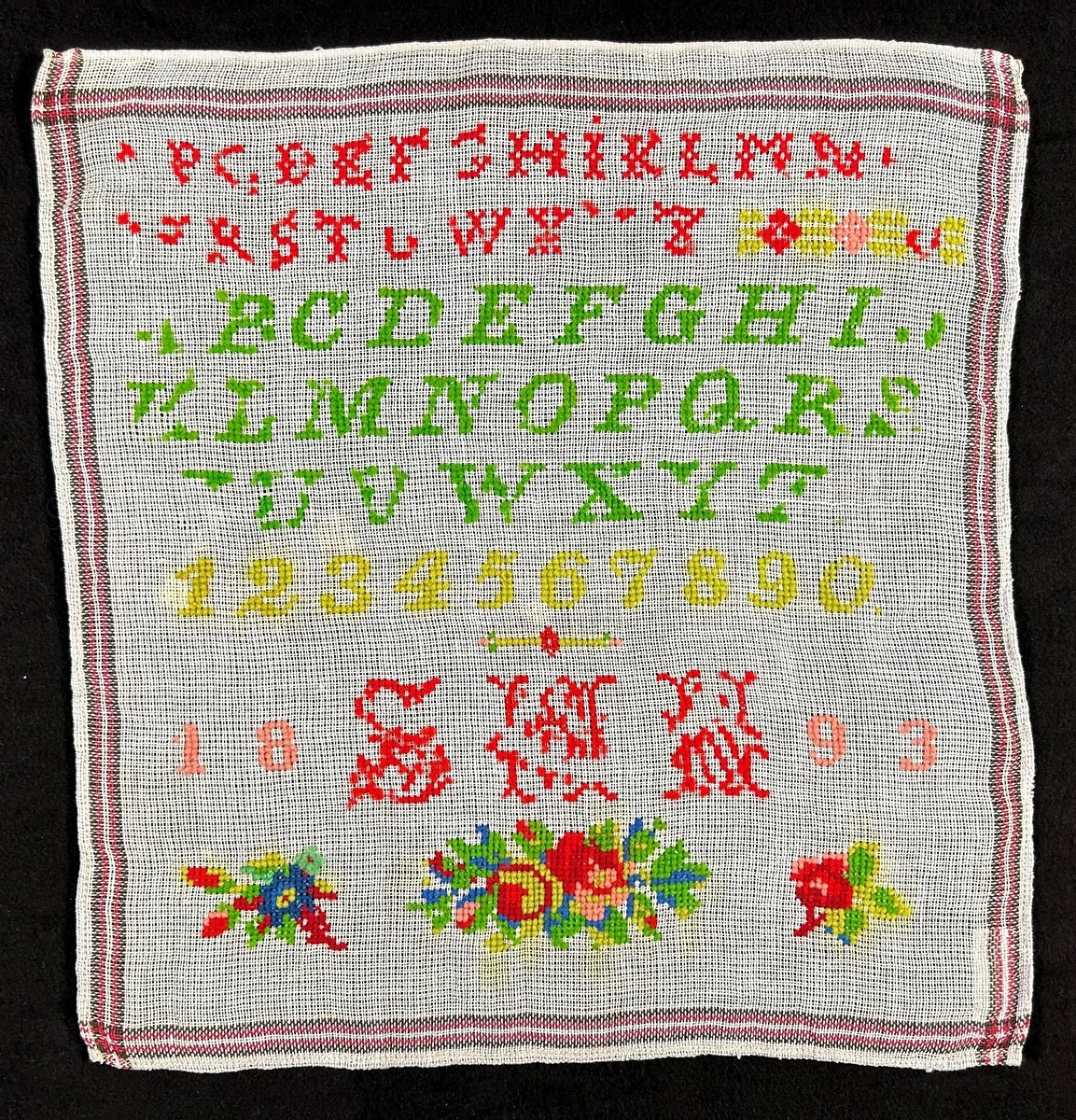 Märkduk på vit botten med korssömsbroderi i rött och grönt. Alfabetet och siffror broderade i rader, Undertill ett monogram: S.M.N. och årtalet 1893. Längst ned blommor.