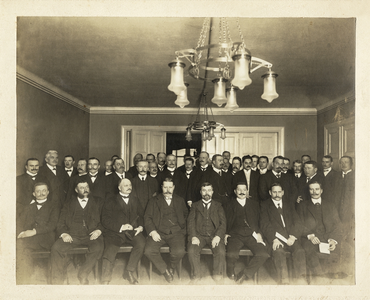 En grupp herrar i mörka kostymer med väst m.m. är avfotograferade i en sal.
På baksidan text: "Foto Sven Wulf, Nybrogatan 8, Stockholm. 
Svenska Arbetsgivareföreningens stora sal, nr 4 fr. vänster SAF:s ordförande Hj. v. Sydow. , andra raden
fr.v. 1) ingenjör Ivar O. Larson, Sthlm. Nr 3 fr. höger med helskägg, direktör Carl Gottwald Fogelberg, Kosta".
