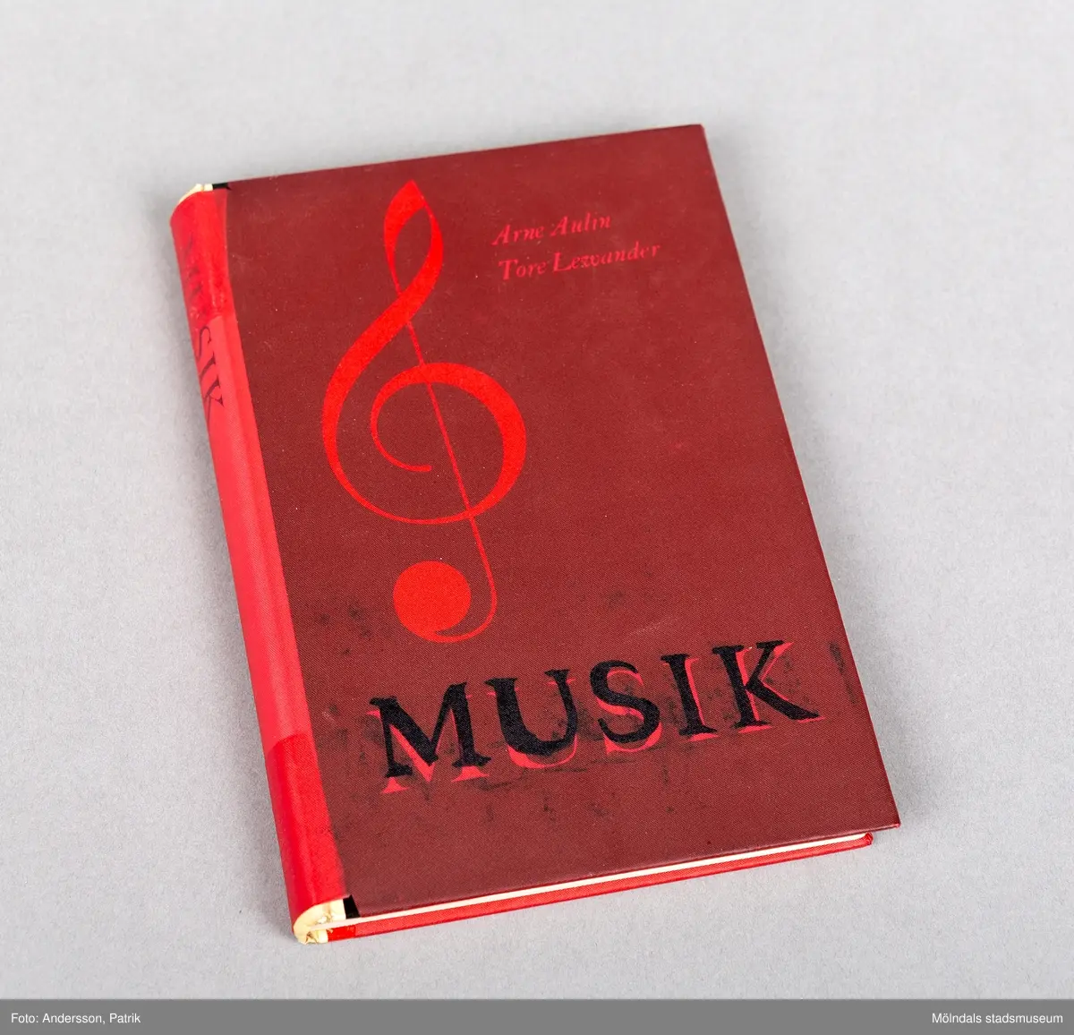 En inbunden sångbok, Framsidan är mörkröd och pryds av en stor not i en ljusare röd färg. 
Längst ned står det Musik med svarta bokstäver. Pärmens rygg är ljusare röd.