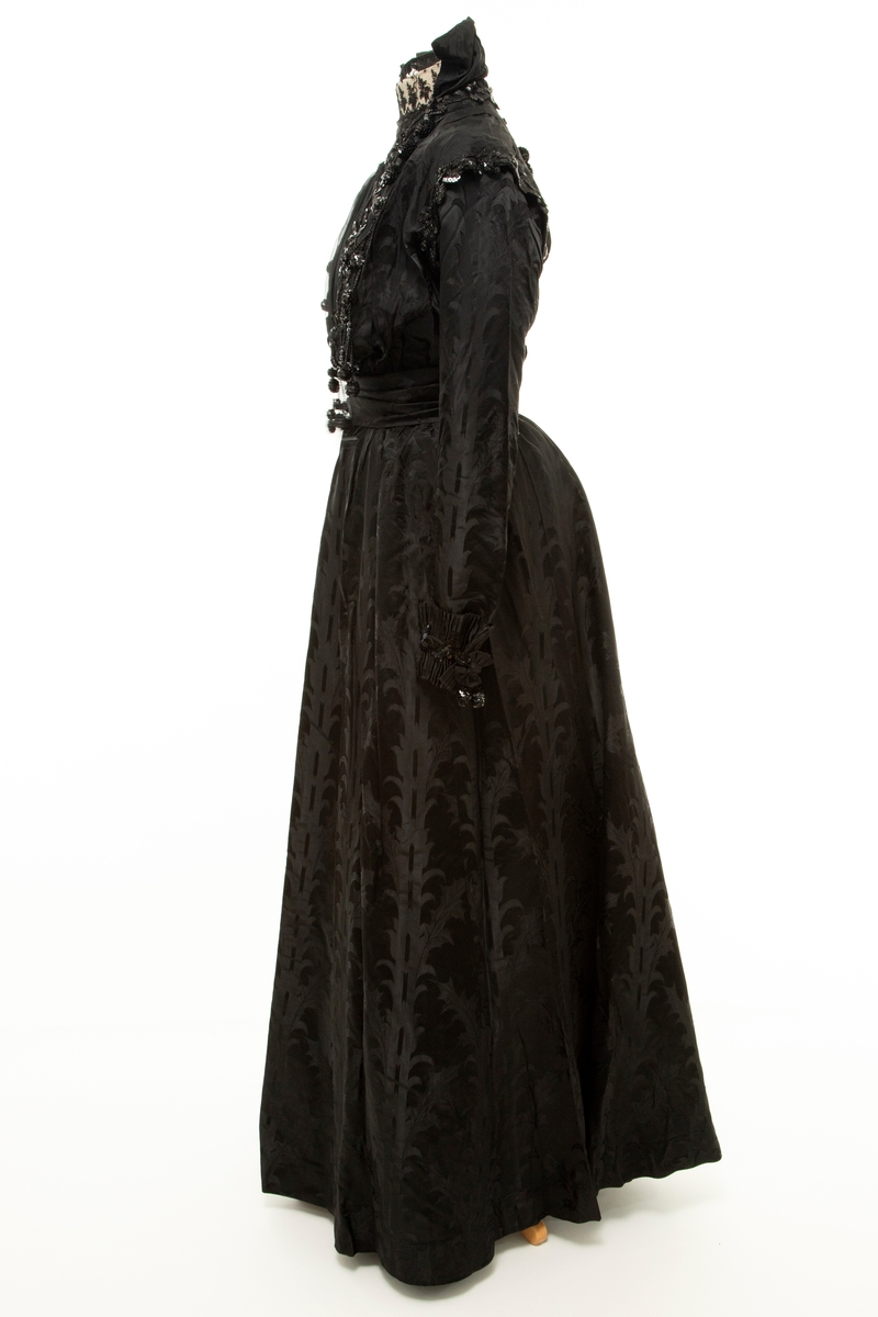 Sortfarget kvinnedrakt i tre deler:

1) Kjoleliv, midjelangt, med frontlukning, lange, trange ermer, delvis løs vest over bysten, chiffon "duebryst" og blonde ståkrage innenfor en Maria Stuart-krage. Mye dekorativ bruk av paljetter og steinkullsperler i broderier og possementeri. Fôret.

2) Skjørt, utskrådd og kilesydd, forsiktig rynket til smal linning, noe mer rynket midt bak, på hver side av lukningen. Lukket med hekter. Ufôret.

3) Belte, drapert og ufôret. Splittet i to i hver ende. Endestykkene lukkes med henholdsvis én og to trykknapper skjult av hver sin sløyfe. Sydd for hånd.