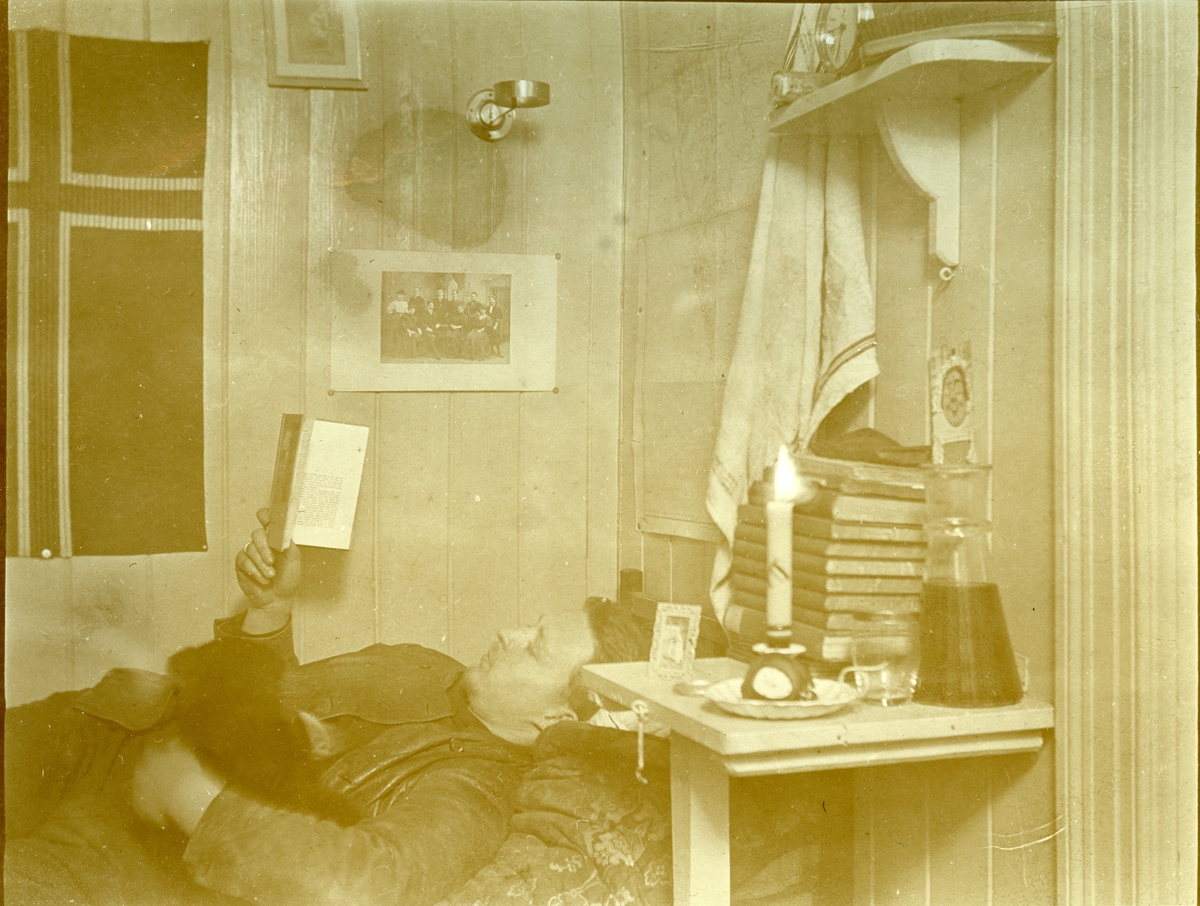Jens Kristoffer Bay på rommet sitt i Longyear City i 1914/15. På baksiden av bildet har han skrevet: Den stilling hvori jeg tilbragte det meste av mørketiden. (Jumbo i armen)  På veggen henger et familiebilde og et flagg.  I 1914-15 var han sjef for en overvintringsekspedisjon på Svalbard. Han var født i 1891 og var bror til Karl A.Bay.