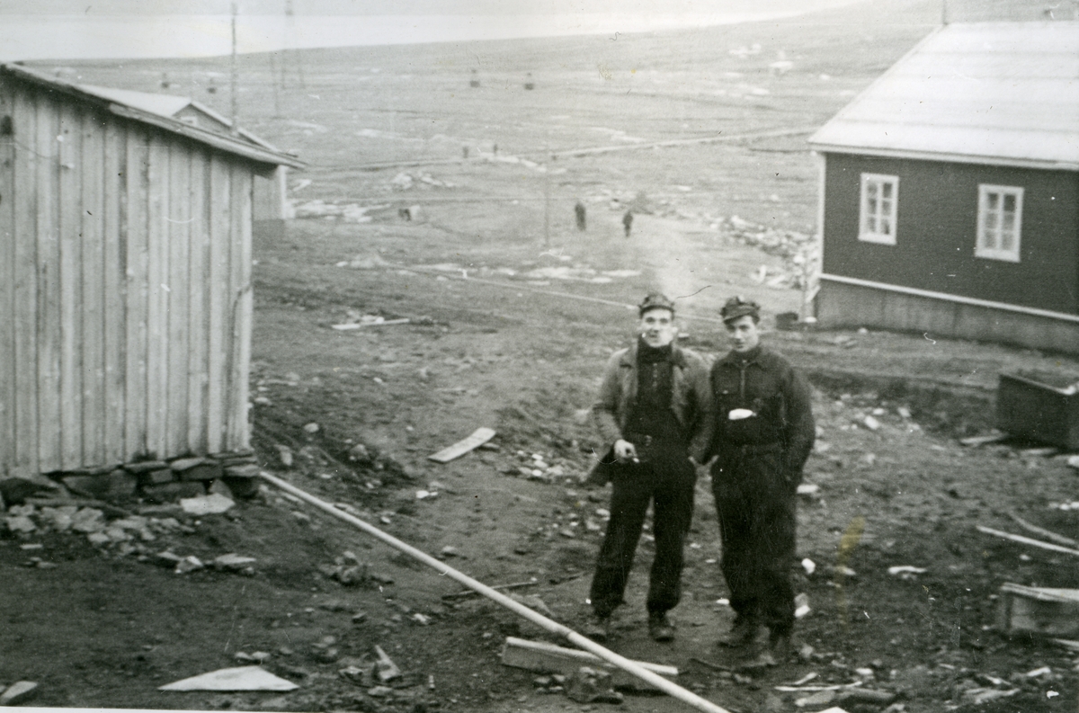 To menn og hus i Longyearbyen i forgrunnen. Flere personer ute på myra. Taubanen med taubanebukker/tårn til  til Gruve 2 a og 2 b i 1940/41. Det er bygd en gangbro over elva mellom byen og Sukkertoppen. Bildet er fra Jens A. Bay sin bildesamling. Han overvintret på Svalbard i 1940/41.