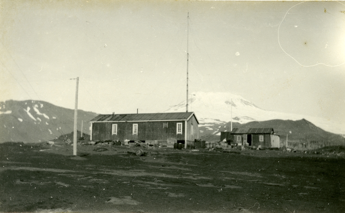 Bilde fra samling etter Emanuel Fredrik Hagbarth Winge, f. 01.10.1904, d. 1956. Han var tannlege i Longyearbyen somrene 1935 og 36. Hans bror Sverre Winge var lege i samme tidsrom, men han overvintret. Originalene er returnert til  eier. Stasjonen og maskinhus på Jan Mayen sett fra SV, Beereberg (2545 moh) bak.