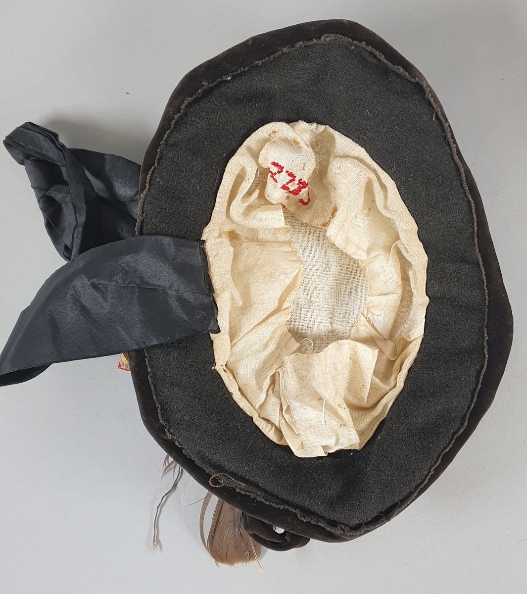 Svart, hjemmesydd hatt til pike. Sydd av ull, med flat pull og smal brem, med flettet fløyelsbånd rundt pullen. Fjær og sløyfe bak på høyre side. Svart sløyfe til å knyte under haken, sløyfen er falt av på den ene siden. Fôret med lin og strie.