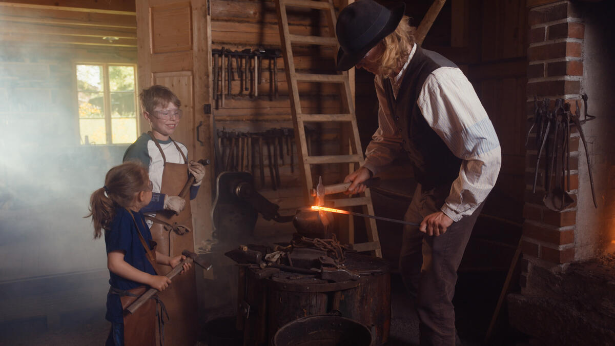 En smed i historisk drakt som arbeider med glødende jern over en ambolt i en tradisjonell smie.