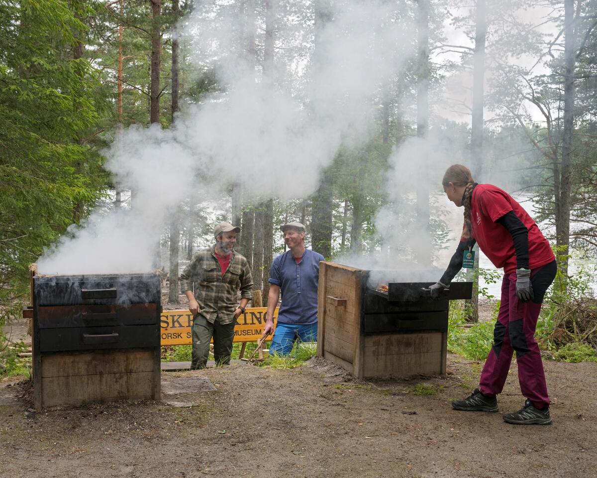 Tilbereding av varmrøykt røye under De Nordiske Jakt- og Fiskedagene 2023 på Anno Norsk Skogmuseum, Elverum, Innlandet. Fiskerøyking, varmrøykingsanlegg for fisk på Prestøya. Ida Skoglund sjekker ei nettingramme med varmrøkt røye. Erik Burud og Håkon Grindal i bakgrunnen. Røyking av fisk.