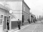 Två män står utanför en Urhandel på Brahegatan i Gränna. En skylt i form av ett fickur hänger ovanför butikens entré. En vägarbetare och några barn längre bort på gatan. En fastighet skyltar för likkistor.
Gatan är belagd med kullersten och utmed fastigheterna till vänster löper två rader med så kallad borgmästarsten utgörande trottoar.