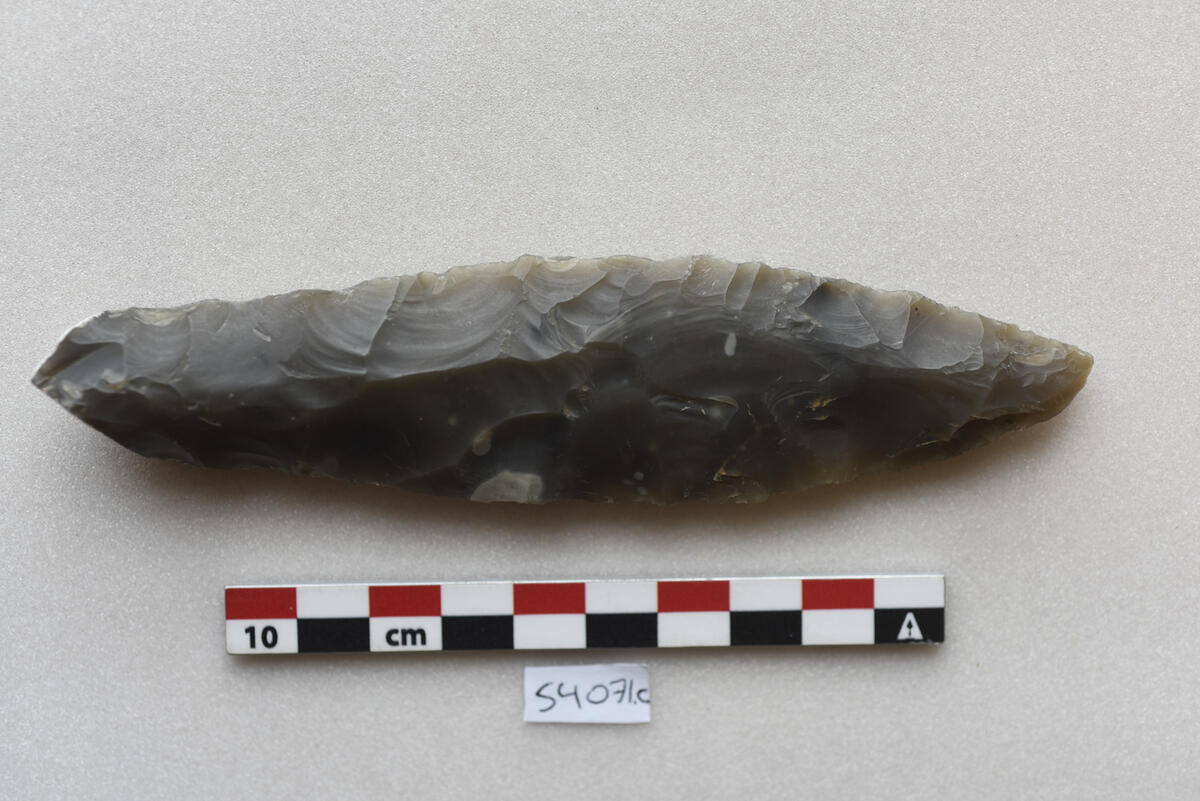 "4071 c. Kortere dolk av graa flint med en del av kalkskorpen sittende igjen i nedre ende og uten særlig skaftdel. Denne form er mere overensstemmende med fig. 8, s. 138 i Nordiske Fortidsminder I, dog er den ikke fuldt saa smal i nedre del som denne fig. Dette stykke er ogsaa smukt arbeidet. Længde 14,3 cm. og bredden 3,4 cm."
Dolk av mørk grå flint, Lomborgs type IIa, i samsvar med beskrivelsen, med cortex på grependen. Stl: 143 mm, stbr: 34 mm, grep stbr: 23 mm, stt: 13 mm.