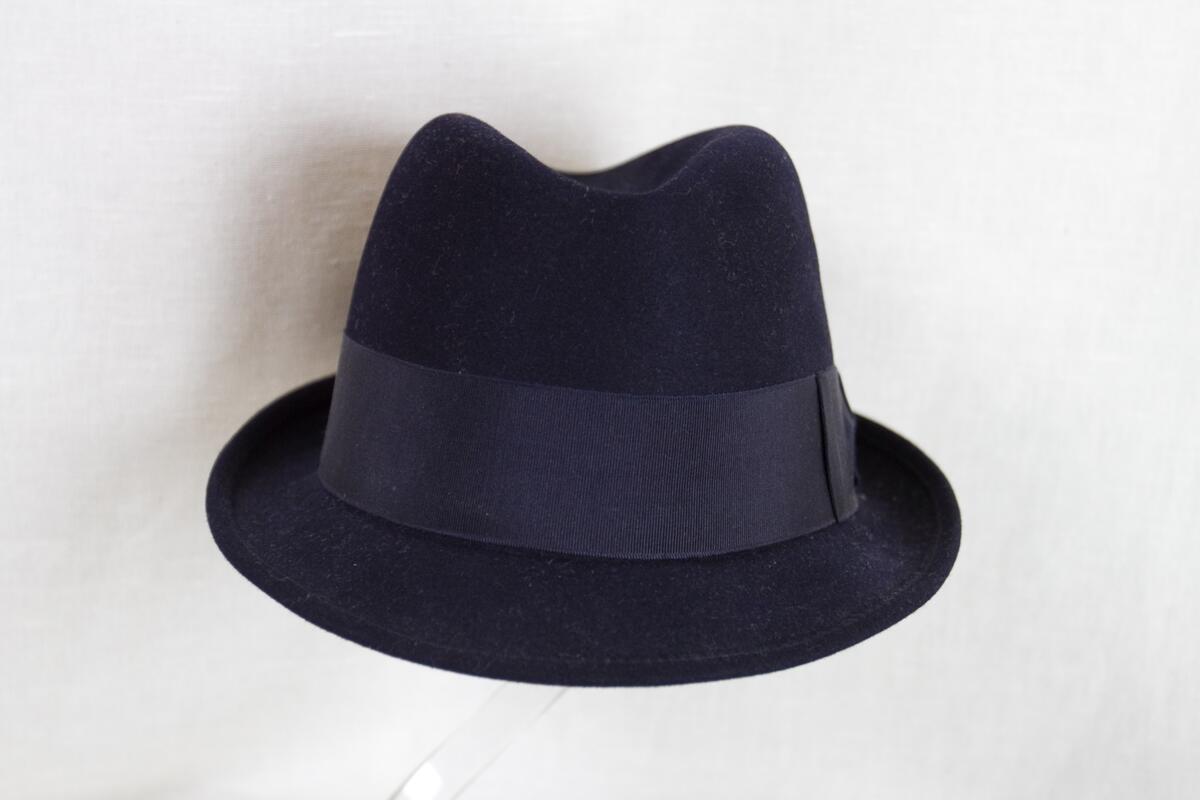 Herrhatt av blå filt. Låg kulle, kraftigt stukad upptill. Smalt brätte, uppvikt bak. 
Mått: Runt kullen 530 mm. Kullens höjd 115 mm. 
Storlek: 55.