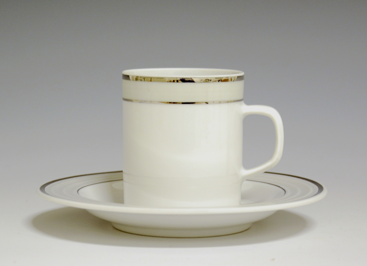 Kopp av porselen med hvit glasur. Dekorert med to enkle striper i platina med et grått bånd i mellom øverst på korpus.
Modell: Saturn
Dekor: Platina
