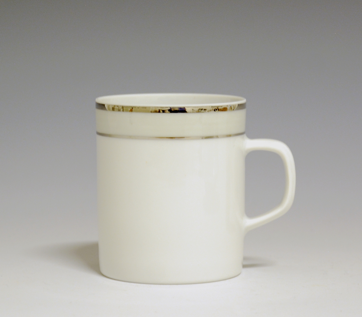 Kopp av porselen med hvit glasur. Dekorert med to enkle striper i platina med et grått bånd i mellom øverst på korpus.
Modell: Saturn
Dekor: Platina
