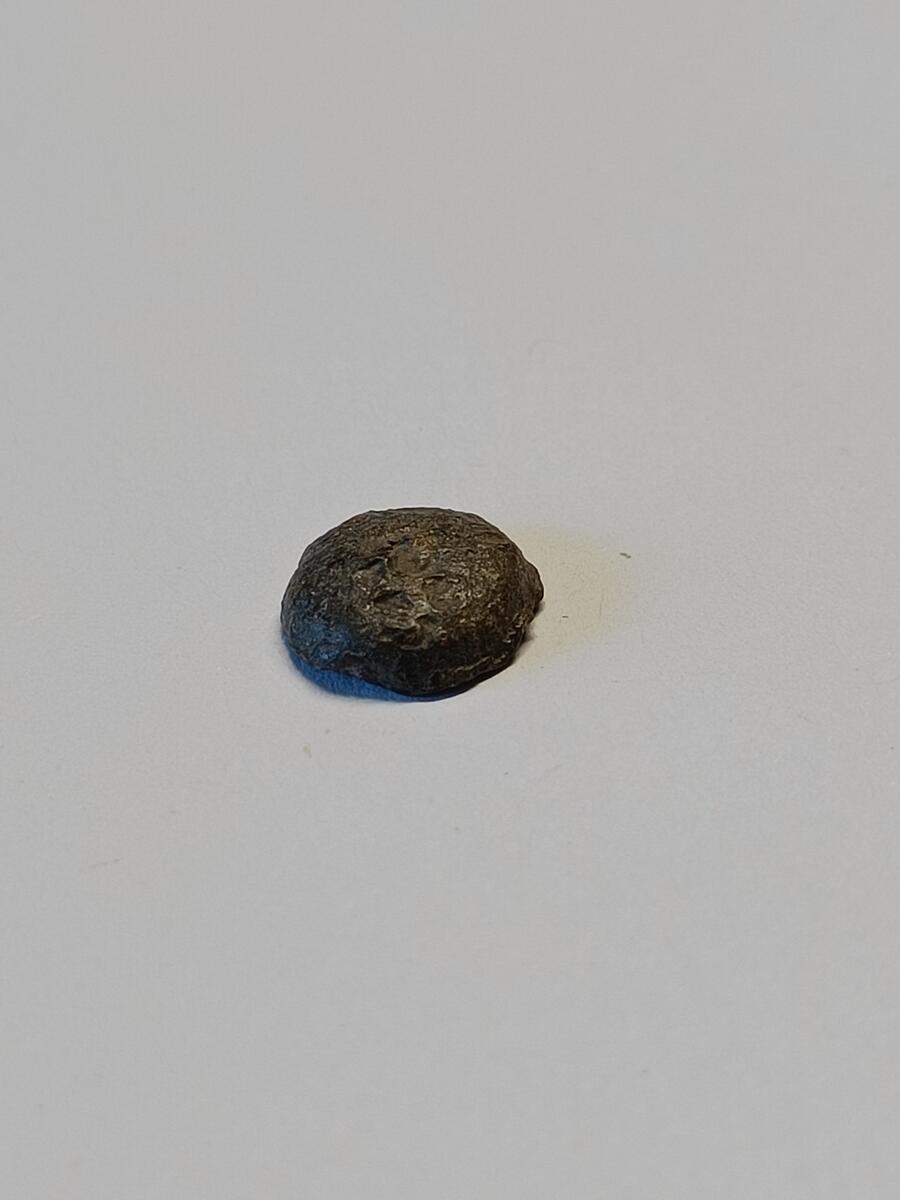 En rund og halvsirkelformet gjenstand, trolig hodet på en knapp. Innfestning til knappens bøyle sees på baksiden, men kun knappehodet er bevart. Grov overflate med et slags hakk i midten av oversiden.