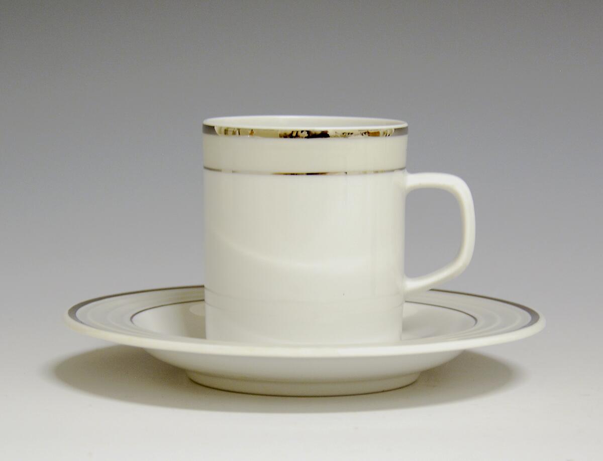 Kopp av porselen med hvit glasur. Dekorert med to enkle striper i platina med et grått bånd i mellom øverst på korpus.
Modell: Saturn
Dekor: Platina
