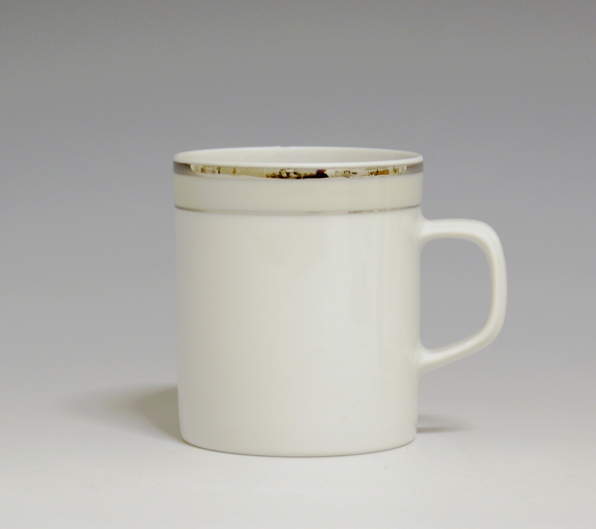 Kopp av porselen med hvit glasur. Dekorert med to enkle striper i platina med et grått bånd i mellom øverst på korpus.
Modell: Saturn
Dekor: Platina
