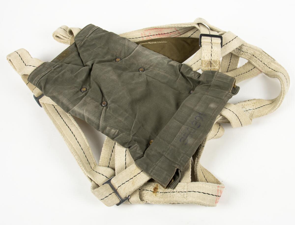 Bröstfallskärm, FASK 22. G.Q, Typ C. Mk.2.
Remmarnas utlösningsmekanism är helt mekanisk. Hölje av smärtingduk. Vit kalott av nylon. Skärmen är utlöst och stämplad Kasserad.
Medföljande tillhörande sele med remmar av sadelgjord.