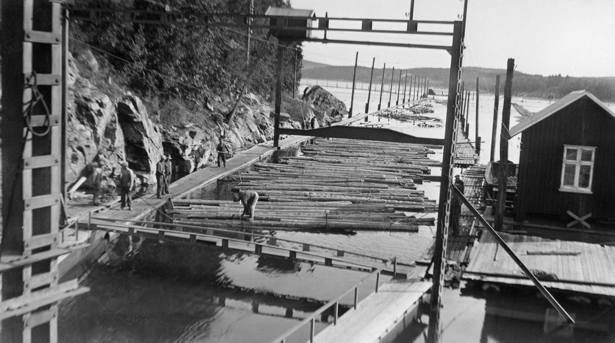 Fra Svartdal eller «Toppen» – inntaket til tømmerkanalen som førte tømmeret i Glommas vestre løp fra Isnesfjorden til Visterflo uten å måtte fløte det via Sølvstufossen.  Fotografiet er tatt fra det punktet der tømmerkanalen gikk inn i fjellet, med kameraet vendt mot inntaksrenna.  I forgrunnen ser vi to digre stålportaler, som antakelig ble brukt til å justere posisjonen til «inntaksskuffen» for å tilpasse den til vannstanden i Isnesfjorden og dermed oppnå en høvelig vannføring inne i tømmertunnelen.  På toppen den bakre av disse stålportalene er det bygd et lite bordskur med saltak og bordvegger, antakelig for å skjerme motoren som ble brukt ved heving og senking av inntaksskuffen. Da dette fotografiet ble tatt sto en mann på tømmeret ute i renna mens en del arbeidskamerater fulgte med fra flåtegangene på begge sider av innløpet. Til høyre i bildet ligger det ei kvilebrakke, en bordkledd bindingsverkskonstruksjon med saltak og med et krysspostvindu.  Det arbeidet 25-30 mann på Toppen, et mannskap som administrativt sett var underlagt lensebestyreren på Eidet.