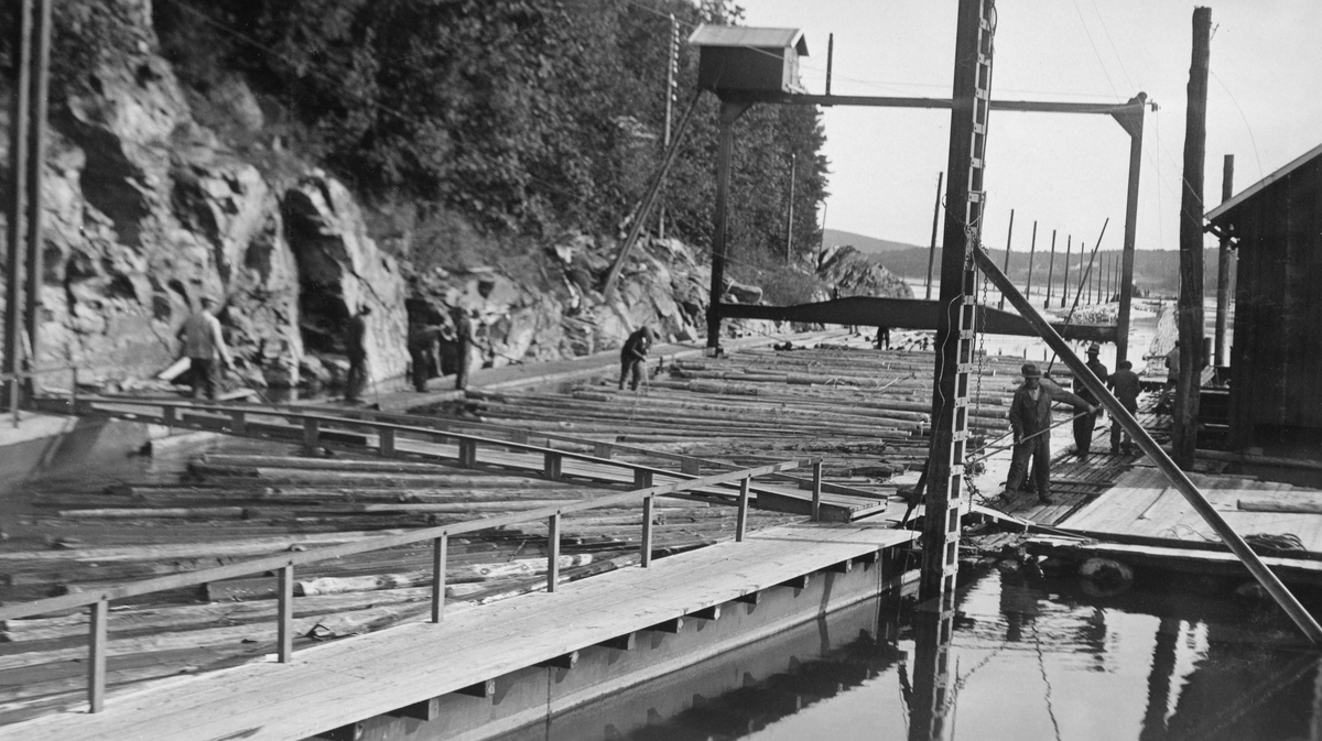 Fra Svartdal eller «Toppen» – inntaket til tømmerkanalen som førte tømmeret i Glommas vestre løp fra Isnesfjorden til Visterflo uten å måtte fløte det via Sølvstufossen.  Fotografiet er tatt fra det punktet der tømmerkanalen gikk inn i fjellet, med kameraet vendt mot inntaksrenna.  I forgrunnen ser vi to digre stålportaler, som antakelig ble brukt til å justere posisjonen til «inntaksskuffen» for å tilpasse den til vannstanden i Isnesfjorden og dermed oppnå en høvelig vannføring inne i tømmertunnelen.  På toppen den bakre av disse stålportalene er det bygd et lite bordskur med saltak og bordvegger, antakelig for å skjerme motoren som ble brukt ved heving og senking av inntaksskuffen. Fotografiet viser også hvordan tømmeret i renna vendes slik at det fra å ligge på tvers i strømretningen justeres til å gå parallelt med kanalens fallretning.  Til høyre i bildet ligger det ei kvilebrakke, en bordkledd bindingsverkskonstruksjon med saltak og med et krysspostvindu.  Det arbeidet 25-30 mann på Toppen, et mannskap som administrativt sett var underlagt lensebestyreren på Eidet.