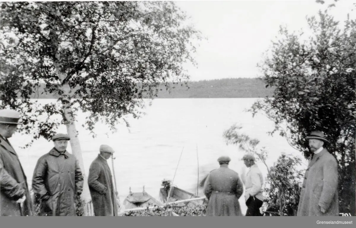 Seks personer står ved vannkanten til Pasvikelven. Noen robåter ligger inntil land. 