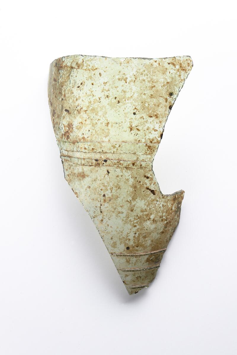 Del av buken på ett ofärgat, kägelformat, optikblåst passglas som är ornerat med fem pålagda parallella trådar. De två övre trådarna är strierade. Fragmetet har glaspest.