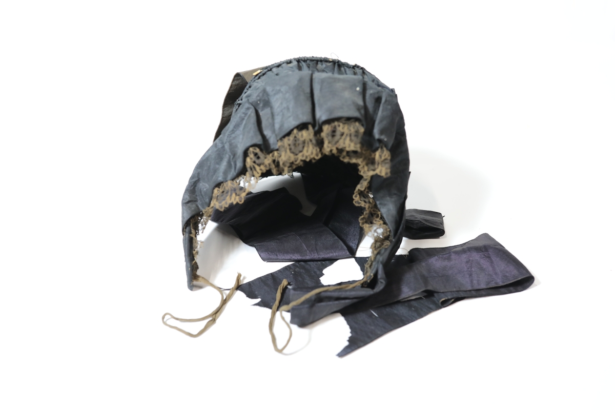 Hette, har form som ei pull-lue med påsydd kappe langs 
nedkant. Sydd av svart silketaft. Sydd inn oppstivande strengar 
av jern i framkant og nedkant, samt 3 stk strengar til langs 
pannestykket, smetta i kanalar og rynka. Pullstykket er også stiva 
opp med fire trespiler. Sydd i kanal og rynka. Innunder framkant 
er påsydd eit brunsvart blondeband lagt i pipefaldar. Samt eit 
taftband, også dette lagt i pipefaldar. I nedkant er påsydd ei 
kappe av same silketaft, lagt i breie faldar. Denne er stiva opp 
med eit lag tyll på innsida. Og i nedkant er påsydd ein svart 
knipling, sydd glatt til nedkant. Pannestykket går ned i djupe 
øyresnippar. Pullstykket er halvmåne-forma. Mellomfor av svart 
stiva kvit bomullsgas. Utanpå denne er påsydd ei brei remse 
brun mugg - bomullslerret. To par knyteband framme i 
øyresnippane. Eit par brune bendelband av bomull, og eit par av 
breitt svart ripsvove silkeband, til knyting utanpå. Desse endane 
er lange, og sløyfa heng ned framme i hals. I overgangen 
mellom kappe og lue er påsydd eit smalt svart fløyelsband. Midt 
oppe på pannestykket er fastsydd ei knytta sløyfe av svart 
silketaft-band. Desse er sydd ned eit lite stykke, og endane heng 
laust ned, like lange som nedkant blonde.