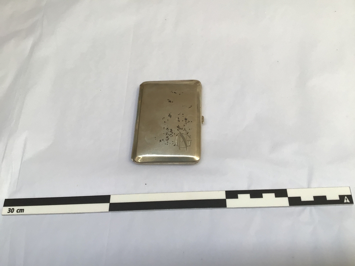 Rektangulært etui med hengsla lok til oppbevaring av sigaretter. Etuiet har initialane til eigar på framsida og ein minnetekst frå givar på baksida.