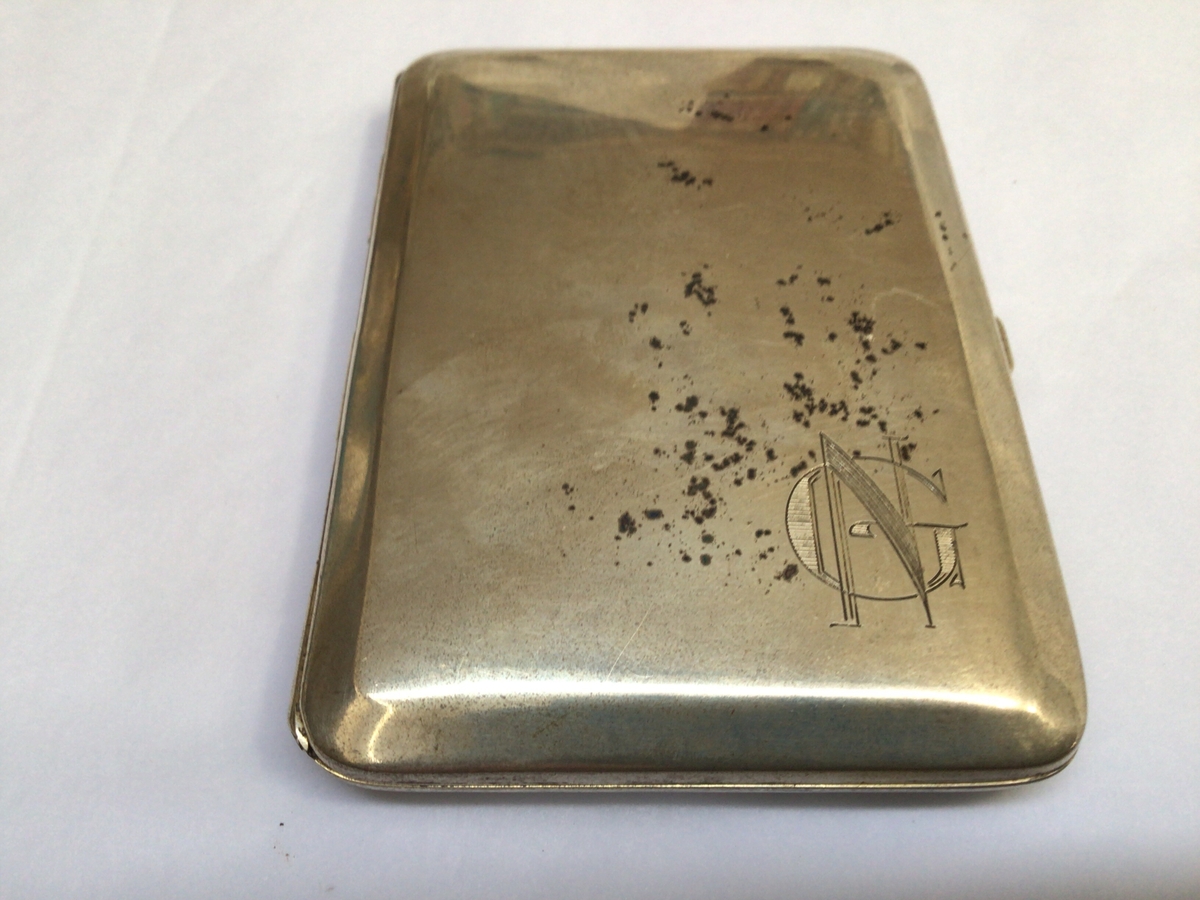 Rektangulært etui med hengsla lok til oppbevaring av sigaretter. Etuiet har initialane til eigar på framsida og ein minnetekst frå givar på baksida.
