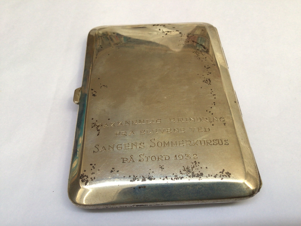 Rektangulært etui med hengsla lok til oppbevaring av sigaretter. Etuiet har initialane til eigar på framsida og ein minnetekst frå givar på baksida.
