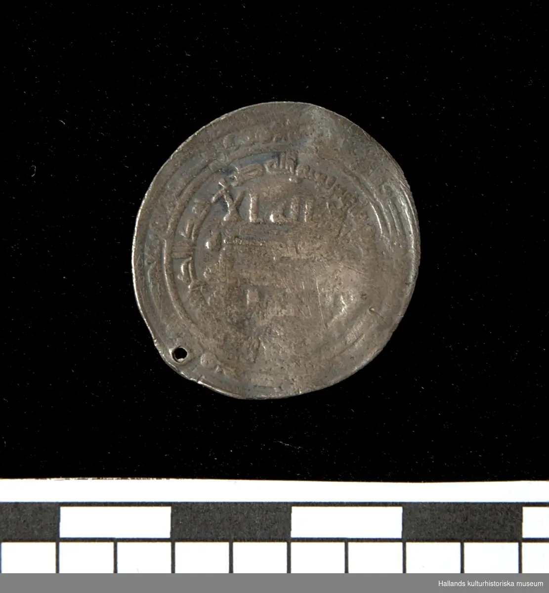 Arabiskt silvermynt (Dirham). Myntet bär ett slaget hål längs kanten. Jordfynd.
