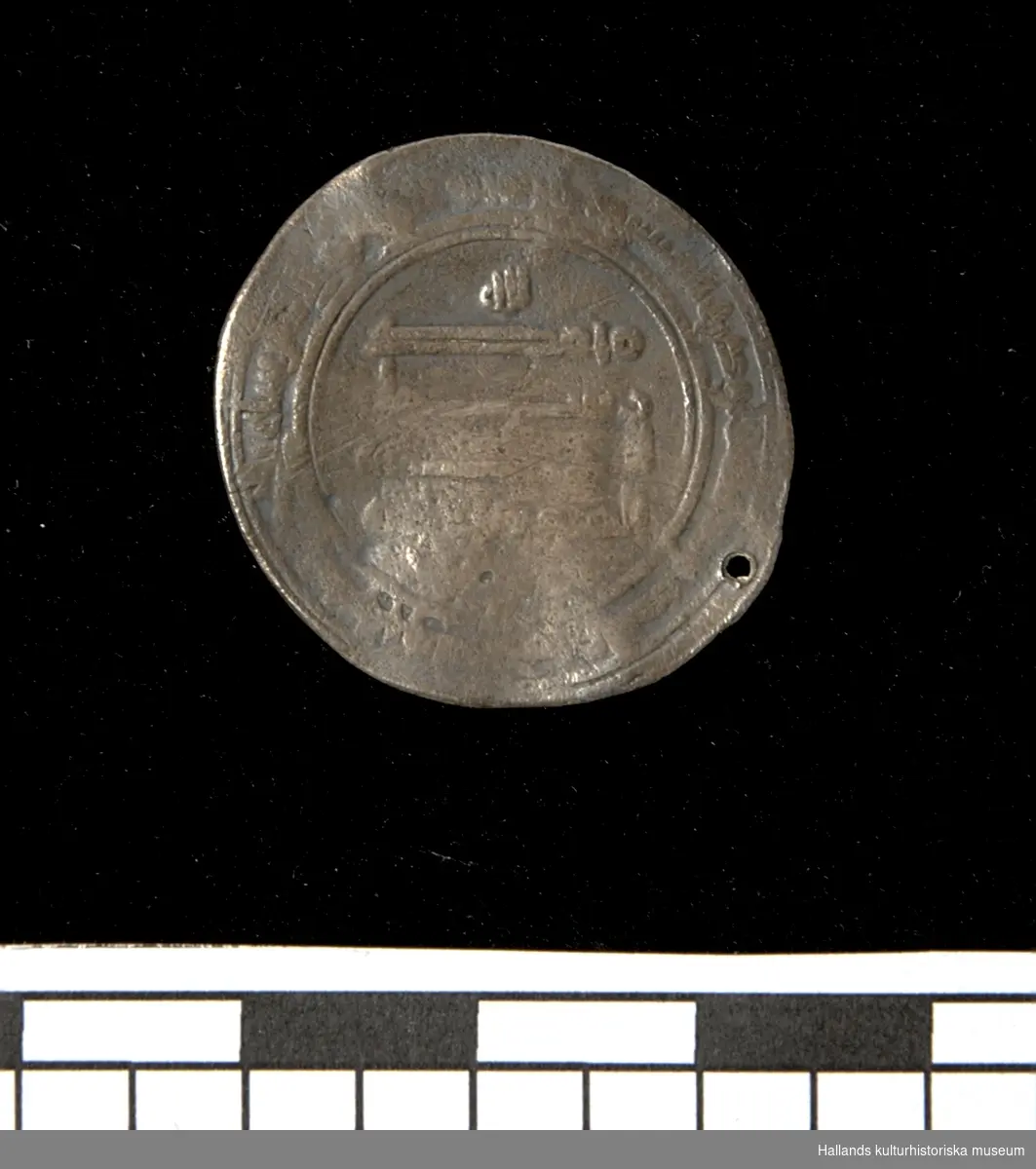 Arabiskt silvermynt (Dirham). Myntet bär ett slaget hål längs kanten. Jordfynd.