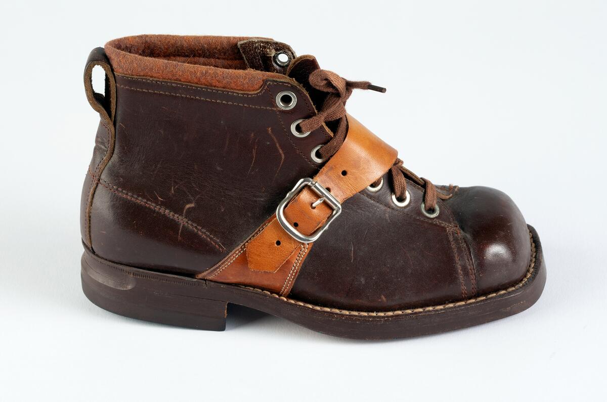 Barnpjäxa av brunt slätt läder. Tvärslå av ljusbrunt läder över vristen med spänne. Sydd och pliggad av ljusbrun lädersula märkt  EKHOLMS. Teknikkänga. Storlek 32. Klacklapp med brunt gummi märkt GOODYEAR. Sju par nitade snörhål. Brunt trikåband. Hälla bak för pådragning. Skåra i klackens baksida. Runt överkanten brun tygskoning.