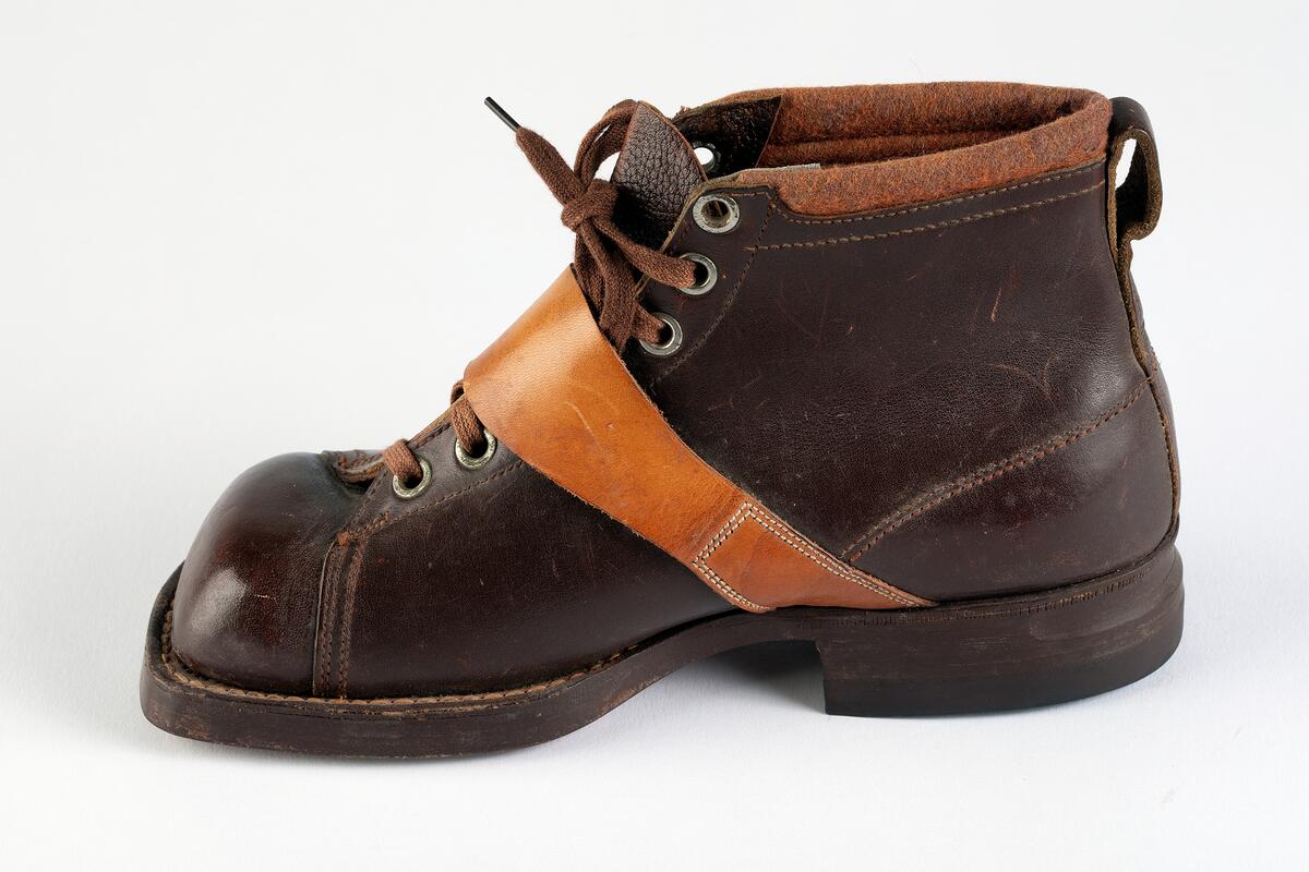 Barnpjäxa av brunt slätt läder. Tvärslå av ljusbrunt läder över vristen med spänne. Sydd och pliggad av ljusbrun lädersula märkt  EKHOLMS. Teknikkänga. Storlek 32. Klacklapp med brunt gummi märkt GOODYEAR. Sju par nitade snörhål. Brunt trikåband. Hälla bak för pådragning. Skåra i klackens baksida. Runt överkanten brun tygskoning.