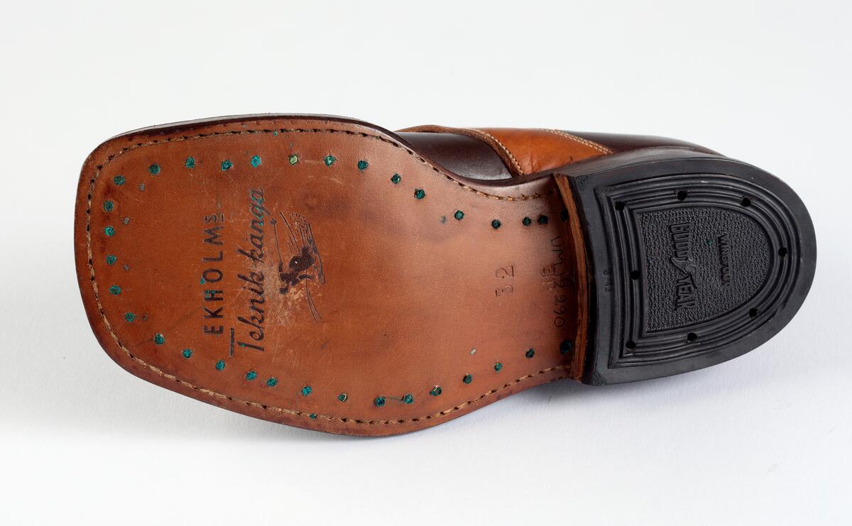 Barnpjäxa av brunt slätt läder. Tvärslå av ljusbrunt läder över vristen med spänne. Sydd och pliggad av ljusbrun lädersula märkt  EKHOLMS. Teknikkänga. Storlek 32. Klacklapp med brunt gummi märkt GOODYEAR. Sju par nitade snörhål. Brunt trikåband. Hälla bak för pådragning. Skåra i klackens baksida. Runt överkanten brun tygskoning.