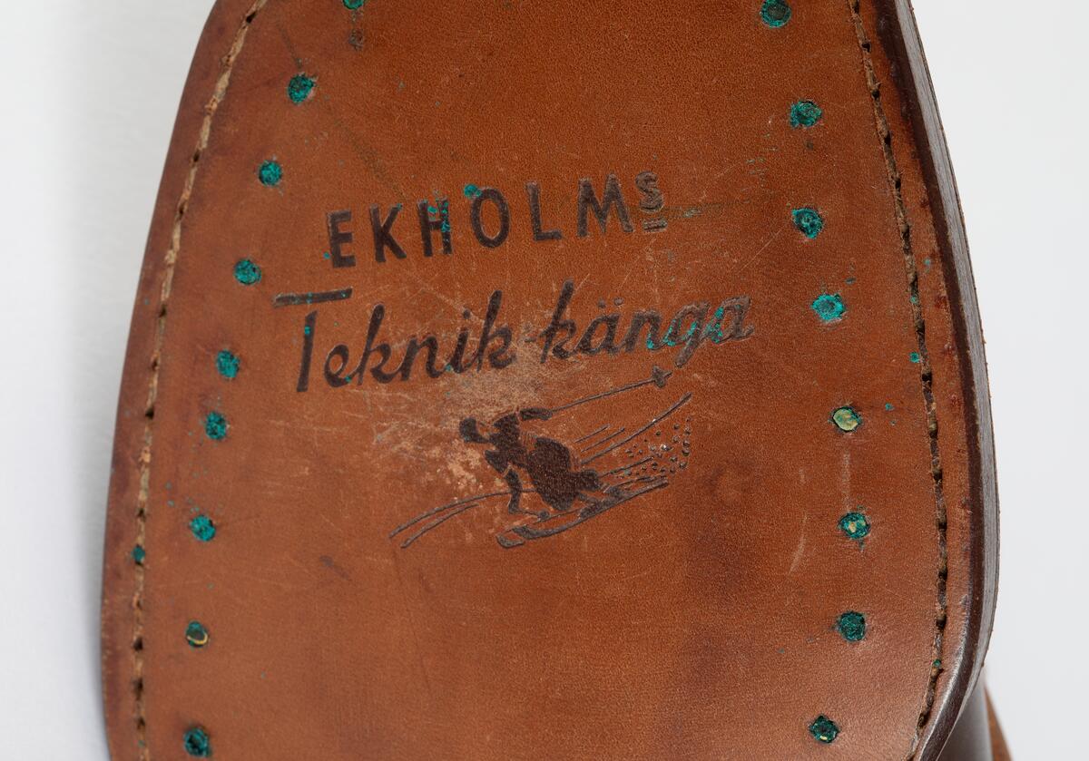 Barnpjäxa av brunt slätt läder. Tvärslå av ljusbrunt läder över vristen med spänne. Sydd och pliggad av ljusbrun lädersula märkt  EKHOLMS. Teknikkänga. Storlek 32. Klacklapp med brunt gummi märkt GOODYEAR. Sju par nitade snörhål. Brunt trikåband. Hälla bak för pådragning. Skåra i klackens baksida. Runt överkanten brun tygskoning.