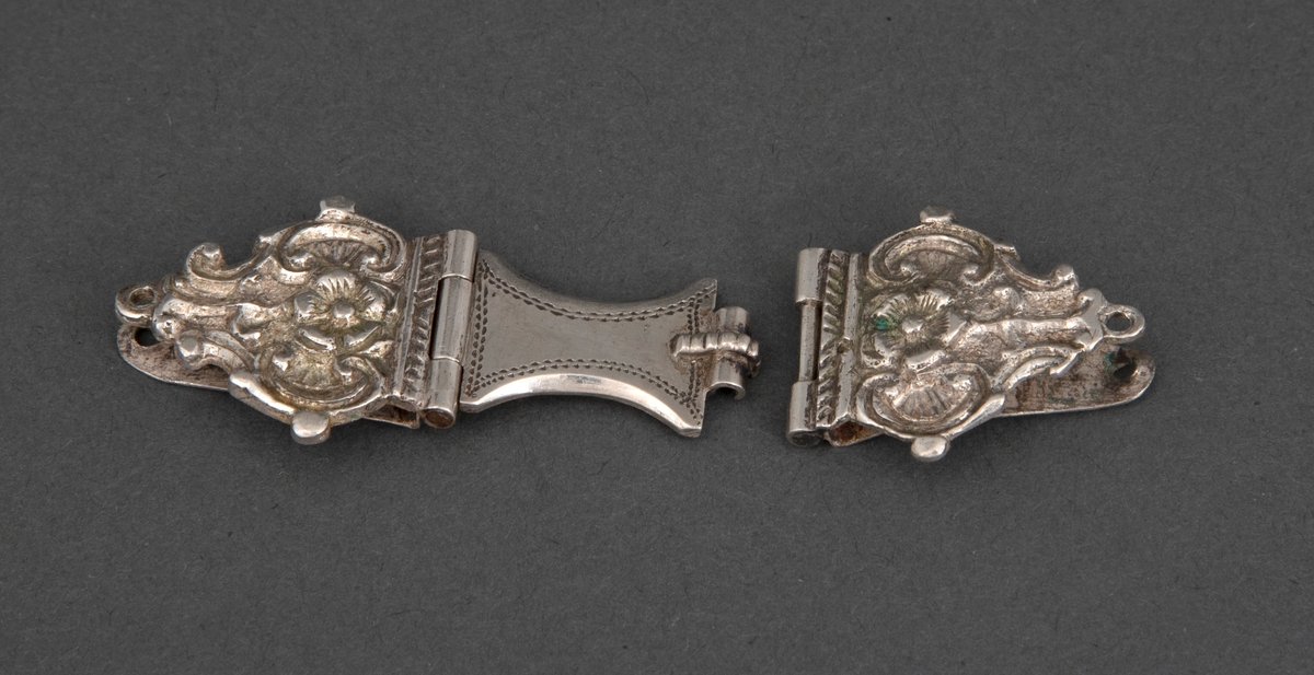 En av to bokspenner i sølv. Spennen er tre-delt og består av to sidestykker og et mellomledd. Mellomleddet er hengslet til det ene sidestykke og griper over en spalte i det andre. Hvert sidestykke består av to plater, som griper om permen. Den ytterste er støpt og siselert, nærmest palmettformet, med C-former og rosett i relieff, samt stifthull. Mellomstykket er glatt med buede langsider og prikkdekor.