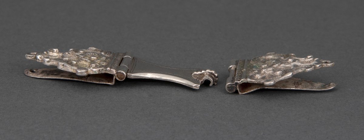 En av to bokspenner i sølv. Spennen er tre-delt og består av to sidestykker og et mellomledd. Mellomleddet er hengslet til det ene sidestykke og griper over en spalte i det andre. Hvert sidestykke består av to plater, som griper om permen. Den ytterste er støpt og siselert, nærmest palmettformet, med C-former og rosett i relieff, samt stifthull. Mellomstykket er glatt med buede langsider og prikkdekor.