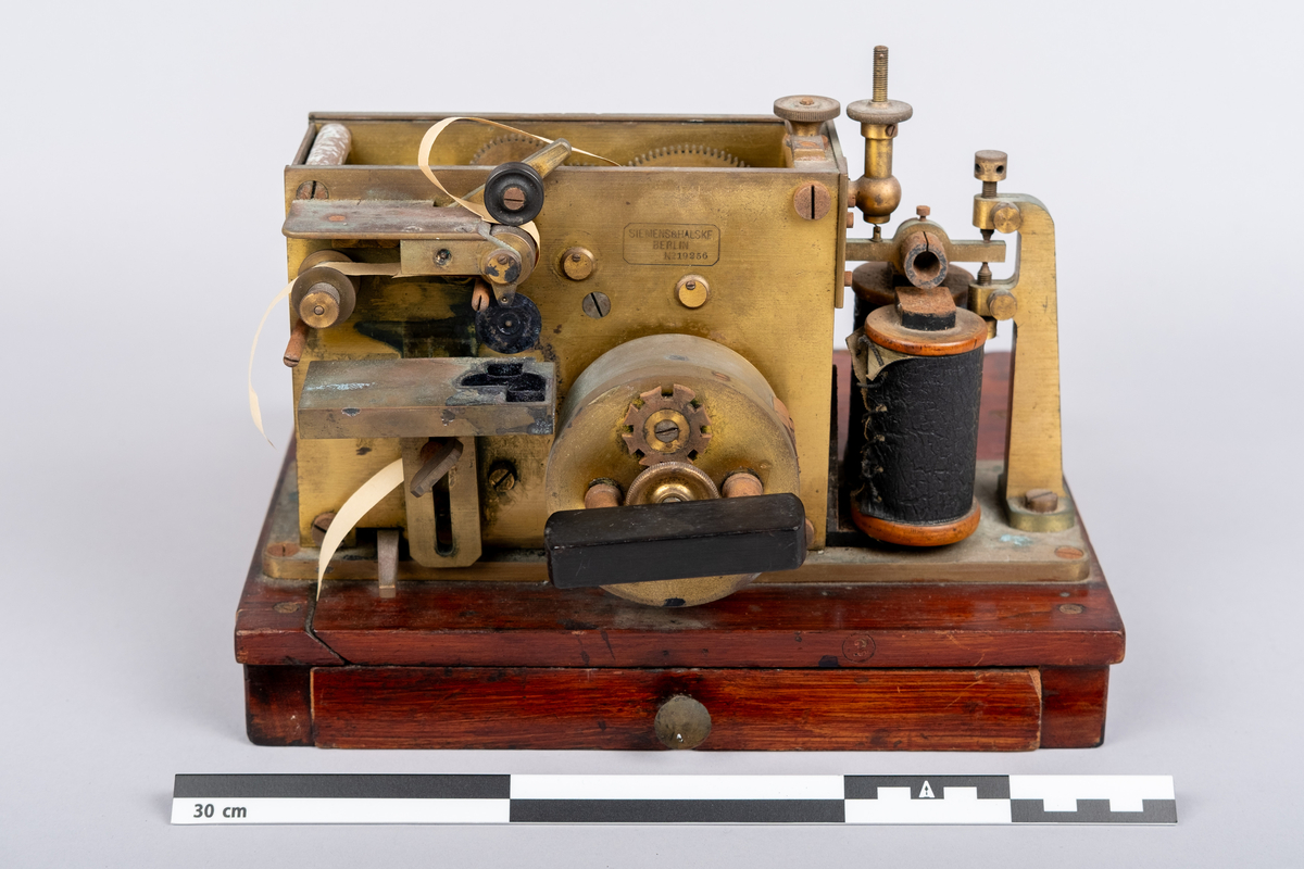 Et morseapparat av typen Siemens & Halske (Berlin) No. 19256. Apparatet har en base av rødmalt treverk og apparatet festet på toppen. Basen har en skuff hvori en papir rull er instarert og innført i apparatet. Basen har på den ene siden et vindu hvor papir rullen kan sees.