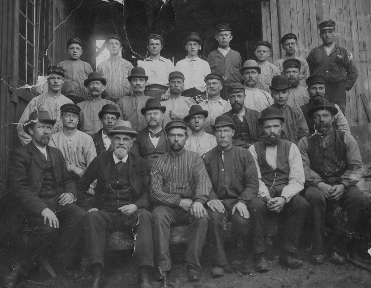 Arbetsstyrkan vid Rylander & Rudolphs Fabriks AB. Övre raden från vänster: A. Vesterberg, J. Andersson, A. Pettersson, V. Stenberg, E. Hultman, F. Karlsson, J. Vesterström samt Moje. Andra raden från vänster: V. Strömkvist, Berggren "Flodhästen", Karlsson "Nordens Kalle", G. Jonsson, O. Nyström, Svar samt Schottenius. Tredje raden från vänster: Andersson "Slipar Anders", K. Stenberg, H. Hultenberg, J. Rudolphs, Andersson "Maskin Anders", Flidner samt J.F. Eklund. Nedre raden från vänster: John Rudolphs, E. Thalén, Fast, Karlsson "Snickar Kalle", Hellberg samt J. Vesterberg. En av de äldsta industrierna i Nacka - Rylander & Rudolphs Fabriks AB - anlade 1889 i området Henriksborg en fabrik för tillverkning av elektrotekniska artiklar, som bl.a. batterier till vevtelefoner, kolspetsar till båglampor, mikrofonkol och dynamoborstar. Företaget startades av direktör John Rudolphs i Stockholm redan 1882, men fyra år senare flyttade han ut företaget till Nacka kommun, närmare bestämt till Klippan vid Danvikstull - då belägen inom Nacka - och där fick den gamla väderkvarnen tjäna som verkstadslokal. Av utrymmesskäl uppfördes en ny fabrik i tegel 1888. Tegelbyggnaden, som ligger vid Danviks Center ca 200 meter öster om Henriksdals järnvägsstation, står fortfarande kvar. Den svängda fasaden utmed Västra Finnbodavägen har en stark karaktär från sekelskiftet 1900 trots senare tillbyggnader. Byggnaden nyttjas idag som kontorshotell under namnet Henriksborg. Foto 1897.