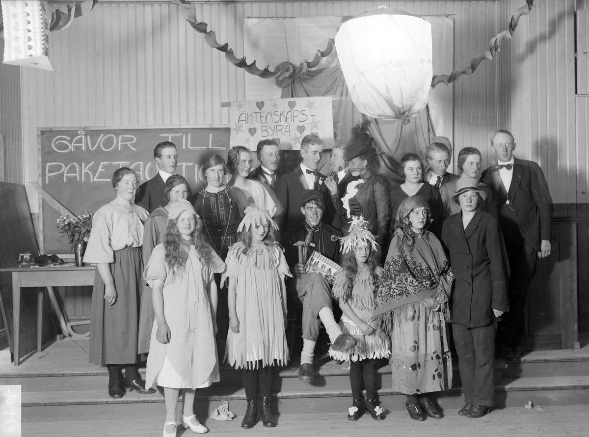 Skolteater i Svinhults kyrkskola 1928. Vi får förmoda att äktenskapliga förvecklingar var ett bärande tema i pjäsen. Bland deltagarna har Iris André i Flinstorp identifierats. Hon står iklädd vit blus alternativt klänning under skylten som annonserar den äktenskapliga byrån.