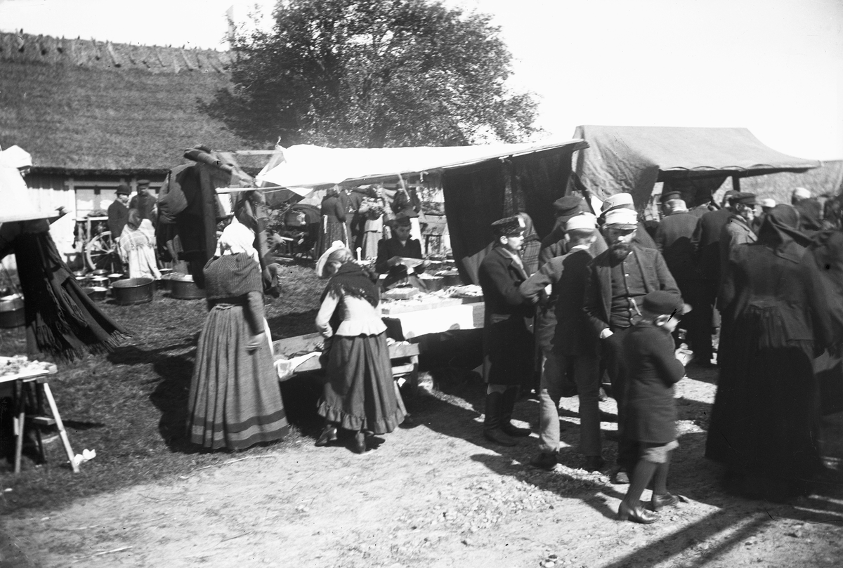 Sjönevads marknad. Marknadsstånd och -besökare vid en byggnad med halmtak. Vid huset säljs kopparkärl av olika storlek.