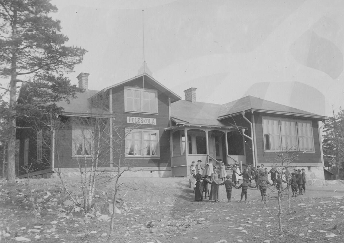 År 1899 uppfördes folkskolebyggnaden Skuru skola. Skolhuset - den äldsta i Skuru - byggdes till 1910 med två nya lärosalar, slöjdsal och två lärarbostäder. Byggnaden revs år 1968. På bilden ses en skolklass som leker en ringlek. Foto 1902.