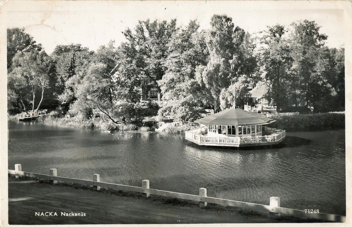 Bilden visar Nackanäs värdshus, men mest dess paviljong ute i vattnet. Fotot är taget från Ältavägenhållet. Nackanäs värdshus verksamhet var igång år 1860-1960, då värdshuset brann upp. Viktor Sjöströms regidebut "Trädgårdsmästaren från 1912" spelades bland annat in på Nackanäs med omnejd. Vykort, 1900-tal. Foto 1900-1960-tal. 