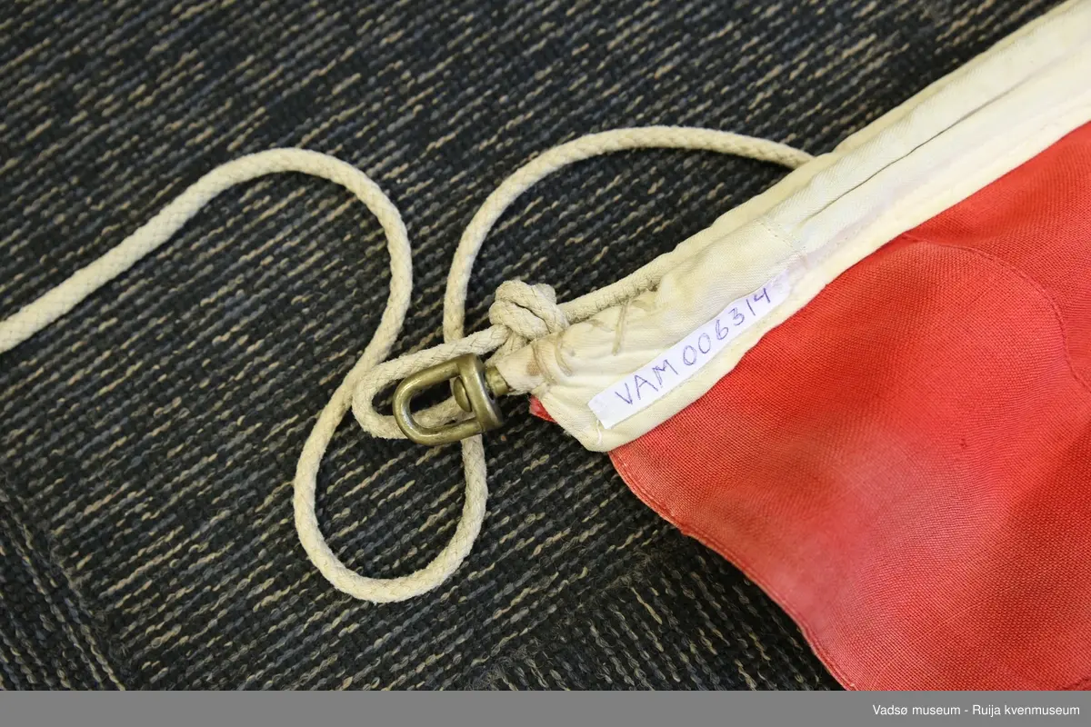 Norsk flagg i vevd materiale, antatt syntetisk. Løpegang og snor av bomullstoff. Flagget har maskinsydd søm, hvitt og rødt er brukt.