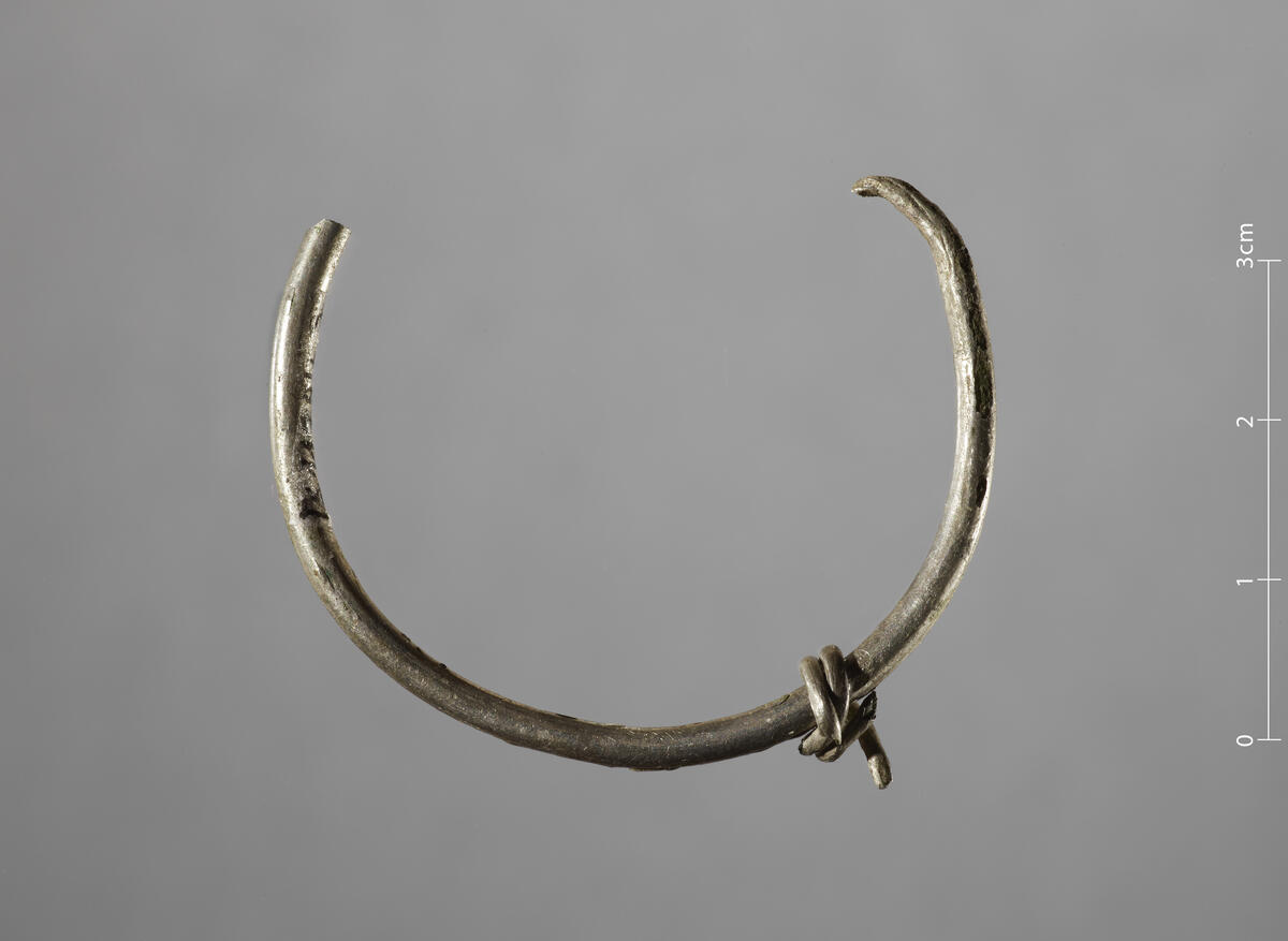 Fragment av sølvring, diam. 4,5 cm, tykkelse 0,25 cm. ¼ av ringen mangler. To tynne spiralfragmenter er festet til ringen.