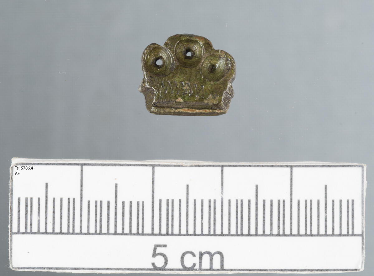 Bestikkende i kobberlegering. Øverst er det "kroneform" i form av tre hvelvinger/ buer. I alle de tre buene er det et lite sirkulært hull midt i buen. Nederst på beslaget er en horisontal linje og over denne dekor i form av horisontal sikksakk-linje. Likner C59577.56 og T20969.170, sant Ts15725, knivendebeslag fra senmiddelalder/nyere tid. 
LokalitetsID: 242090.
Finner: Rikard Larsen