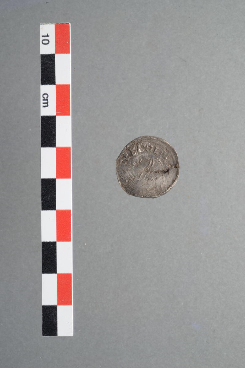 Mynt i sølv. Identifisert av finner som mulig imitasjon av "Long Cross penny" slått under Ethelred (Æthelred, Aethelred) II. Originalen av disse dateres til mellom 997 og 1003 e.Kr. (Skaare 1976 Pl. VII 10-14). Advers har avbildet et mannshode i profil med omskrift rundt. Revers har et likearmet kors i midten med omskrift rundt. Mynten har slitt overflate på begge sider, den er bøyd på midten og delvis sprukket i ene ytterkanten, omskrift er derfor vanskelig å tyde. Se også undernummer 2.
LokalitetsID: 260808