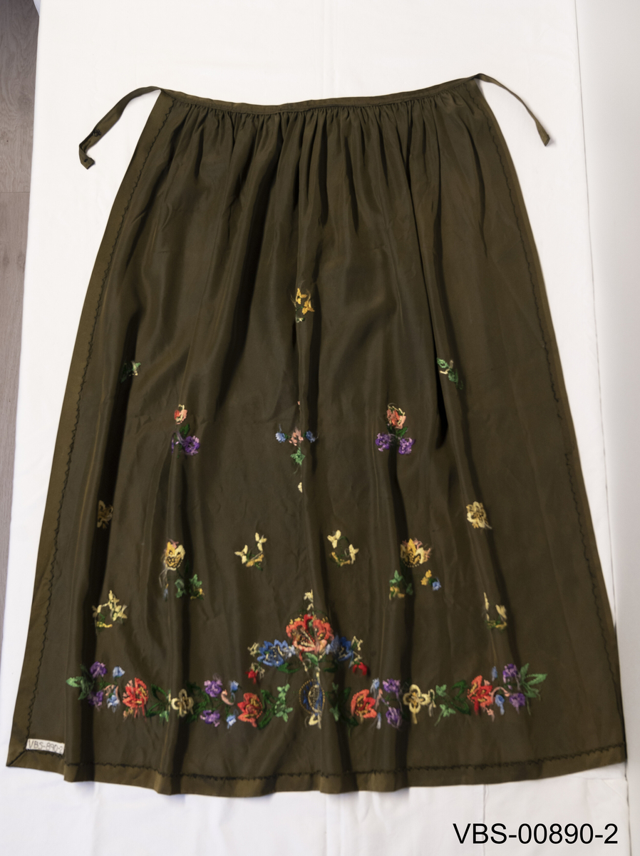 Forkle i glatt tekstil. Grønt med frihåndsbroderi i blomstermotiv.
Sort fløyelsløyfe i midjen (komplement)