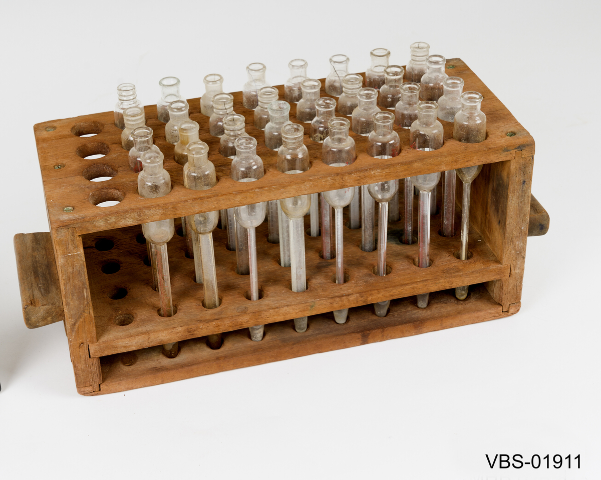 Sett av 32 lignende reagensglass i treholder for bestemmelse av fettinnhold i melk.
Brukte gjenstandene som holder faste og flytende produksjonsrester inne. 
Noen (3) har en gummiplugg midt på halsen på reagensrør.